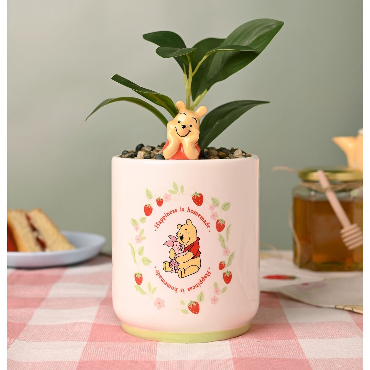 Cream stoneware planter with a 3D Winnie the Pooh figure peeping over the rim, printed strawberry wreath and "Happiness is homemade" design, holding a green leafy plant on a kitchen table.