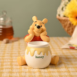 Winnie the Pooh Honey Pot resin storage box, pale yellow bear in red top leaning over a white pot with "hunny" and dripping yellow honey band, shown on a table with honey jar and flowers.