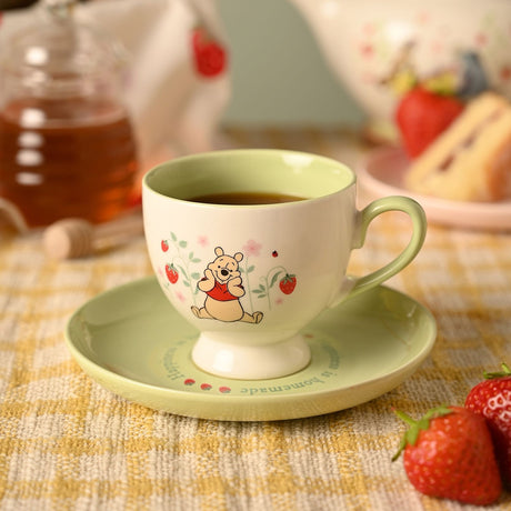 Winnie the Pooh porcelain cup and saucer, 240ml, cream cup with green rim, handle and footed base, Pooh among strawberries and blossoms on a matching green saucer.