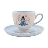 Porcelain footed cup and saucer with Eeyore among pink flowers on a cream cup, pale blue handle and interior, matching blue round saucer, on a white background.