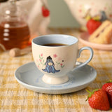 Cream porcelain cup and saucer with pale blue handle and interior, featuring Eeyore among pink flowers and "Enjoy the little things" text, filled with tea beside strawberries.