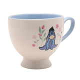 Winnie the Pooh Eeyore porcelain footed cup in cream with pale blue interior and handle, featuring Eeyore among pink flowers and a bee, shown on a white background.