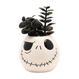 White ceramic planter shaped like Jack Skellington's face with stitched grin, filled with a black faux succulent plant, shown on a plain white background.