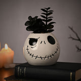 White ceramic Jack Skellington head planter with black stitched grin and eye details, holding a black faux succulent, shown on a book beside lit candles.