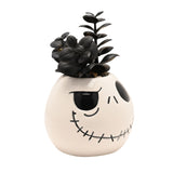 White ceramic planter shaped as Jack Skellington's head with painted black eye sockets and stitched grin, filled with a black faux succulent plant, on a plain white background.
