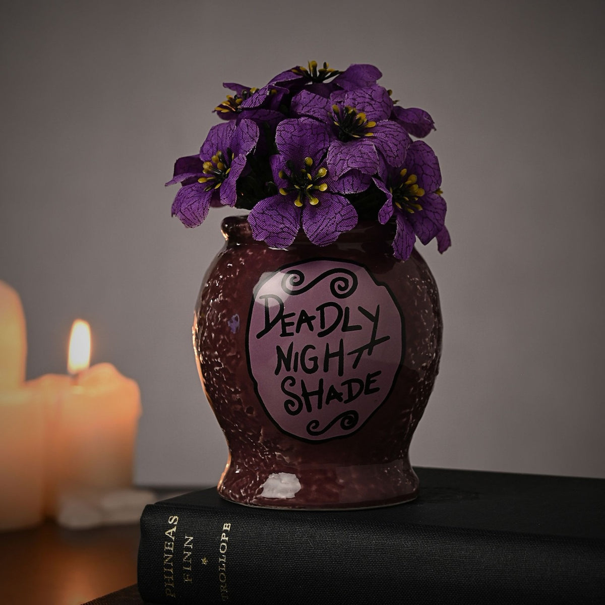 Nightmare Before Christmas Deadly Nightshade Planter in speckled glossy purple with lilac label, holding faux violet flowers, displayed on a book beside candles.