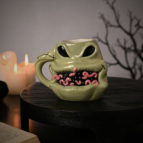 Nightmare Before Christmas Oogie Boogie shaped ceramic mug in sage green, with black eyes, stitched grin, and pink worm detail, shown on a dark table near candles.