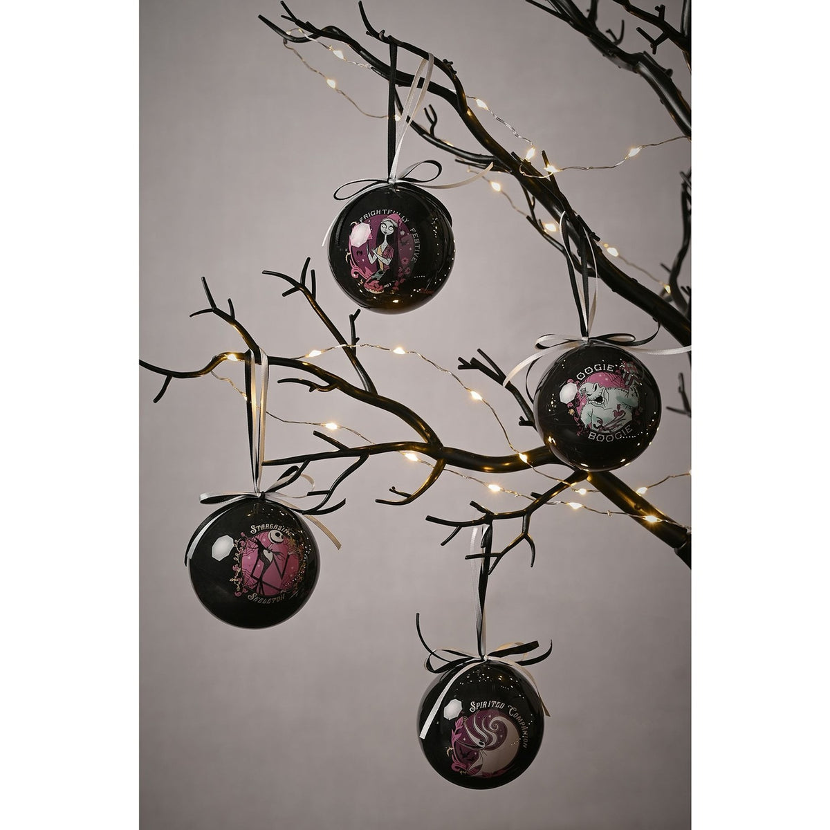 Set of four glossy black round tree baubles hanging from decorative branches with fairy lights, each printed with a pink, white and pale blue Nightmare Before Christmas character panel.