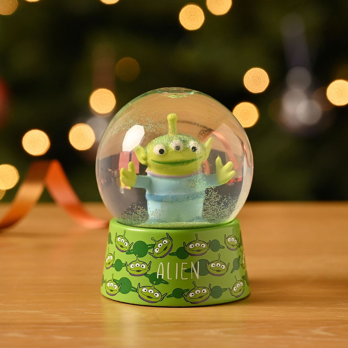 Toy Story Alien Glitter Plastic Snow Globe in clear and green, featuring the three-eyed alien with arms outstretched on a green base printed with alien faces, set against a bokeh light background.