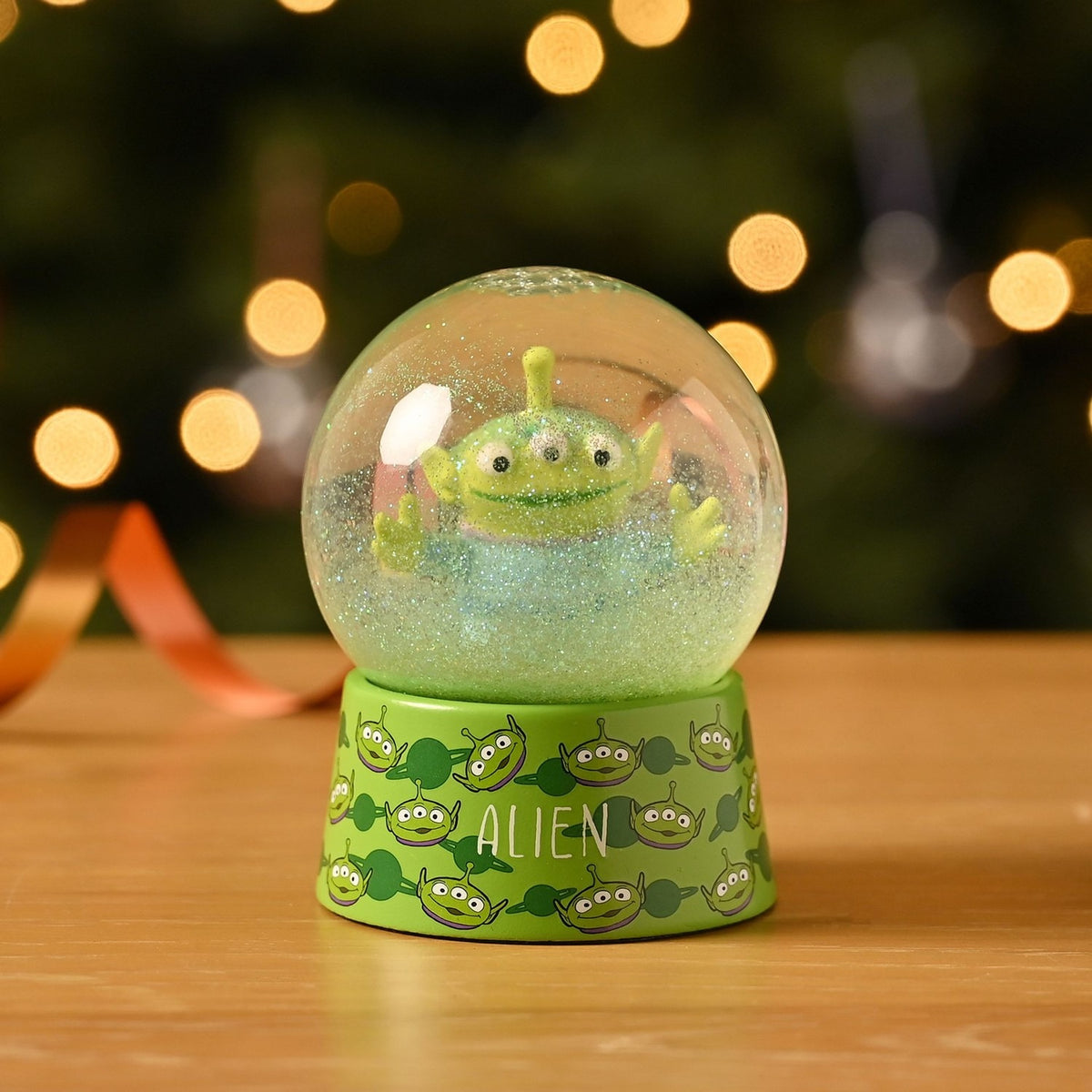 Toy Story Alien Glitter Plastic Snow Globe with green base printed with alien faces, clear dome showing the three-eyed alien figure and green glitter, displayed on a wooden surface.