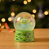 Toy Story Alien Glitter Plastic Snow Globe with green base printed with alien faces, clear dome showing the three-eyed alien figure and green glitter, displayed on a wooden surface.