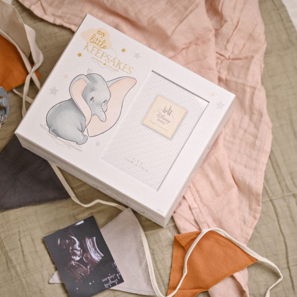 Grey Dumbo Keepsake Box with "my little KEEPSAKES" lettering, Dumbo print, and photo frame window, shown atop pink and olive fabric with dummy clips and a sonogram print.