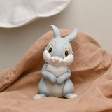 Bambi Thumper Rabbit Money Box in grey, seated with paws folded and ears raised, shown on a pink fabric with a soft neutral background.