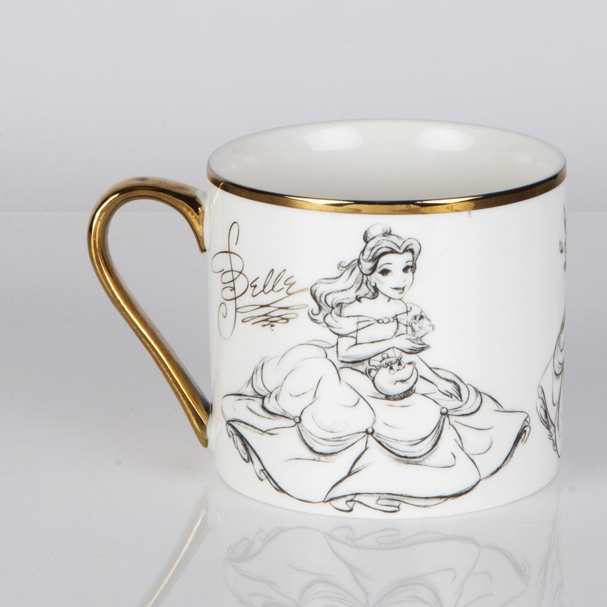 White porcelain mug with gold rim and handle, featuring a pencil-style sketch of Belle in her ball gown holding a teacup, on a plain light background.