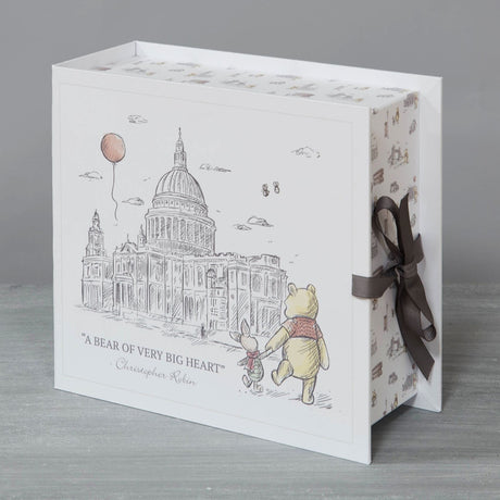 Square cream keepsake box with grey ribbon tie, featuring a sketched cathedral, red balloon, and Pooh bear walking with Piglet above "A Bear of Very Big Heart," shown on a plain grey background.