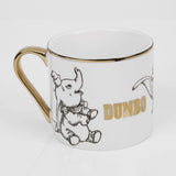 White porcelain Dumbo mug with gold handle and rim, featuring black sketched elephant illustrations and "DUMBO" in gold lettering, shown on a plain white background.