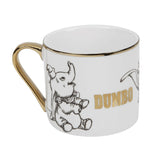 White porcelain Dumbo mug with gold handle and rim, featuring black sketched elephant illustrations and "DUMBO" in gold lettering, shown on a plain white background.