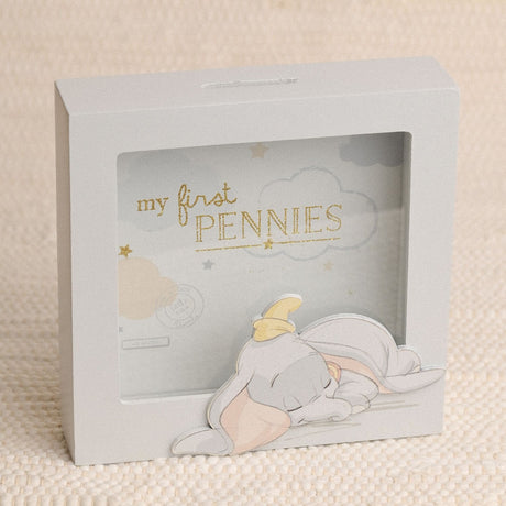 Square grey wooden money box with window front showing a sleeping Dumbo cut-out, pastel clouds, stars, and "my first PENNIES" in gold lettering, on a light background.