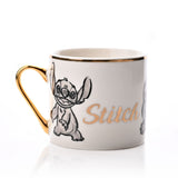 White porcelain mug with gold handle and rim, featuring a grey pencil-style sketch of Stitch and "Stitch" in gold script, set on a plain white background.