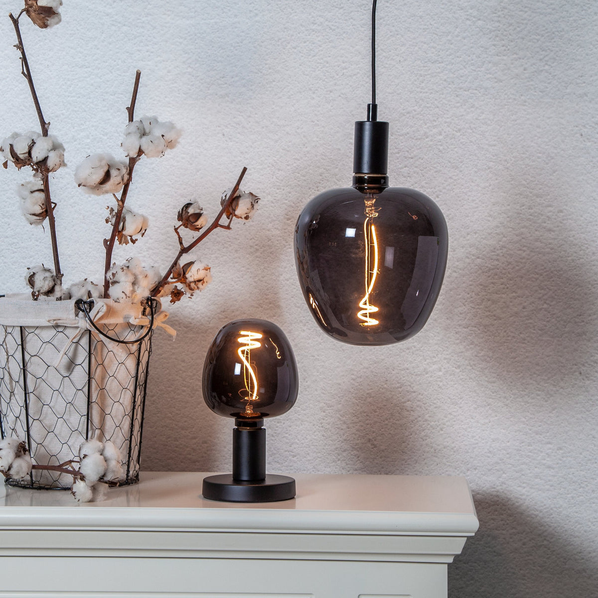 Dimmable LED Bulb E27, smoky black glass with spiral filament, shown lit in a pendant light and table lamp on a cabinet beside a cotton plant display.