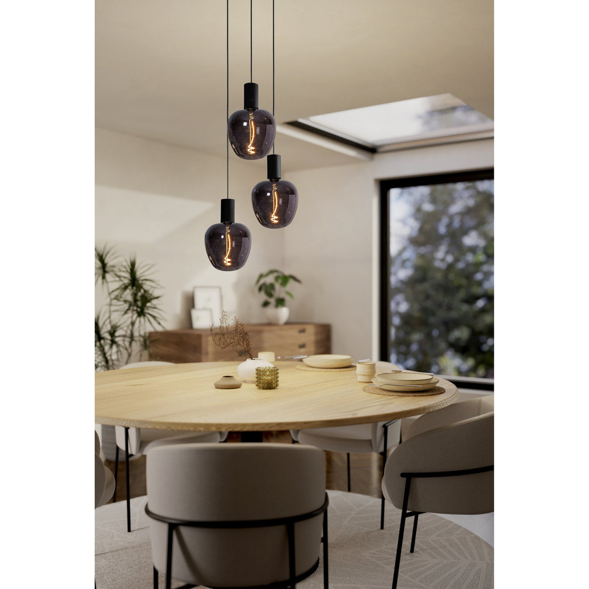 Three smoky black LED pendant bulbs with spiral filaments and E27 screw caps hang over a wooden dining table in a bright room with a skylight.
