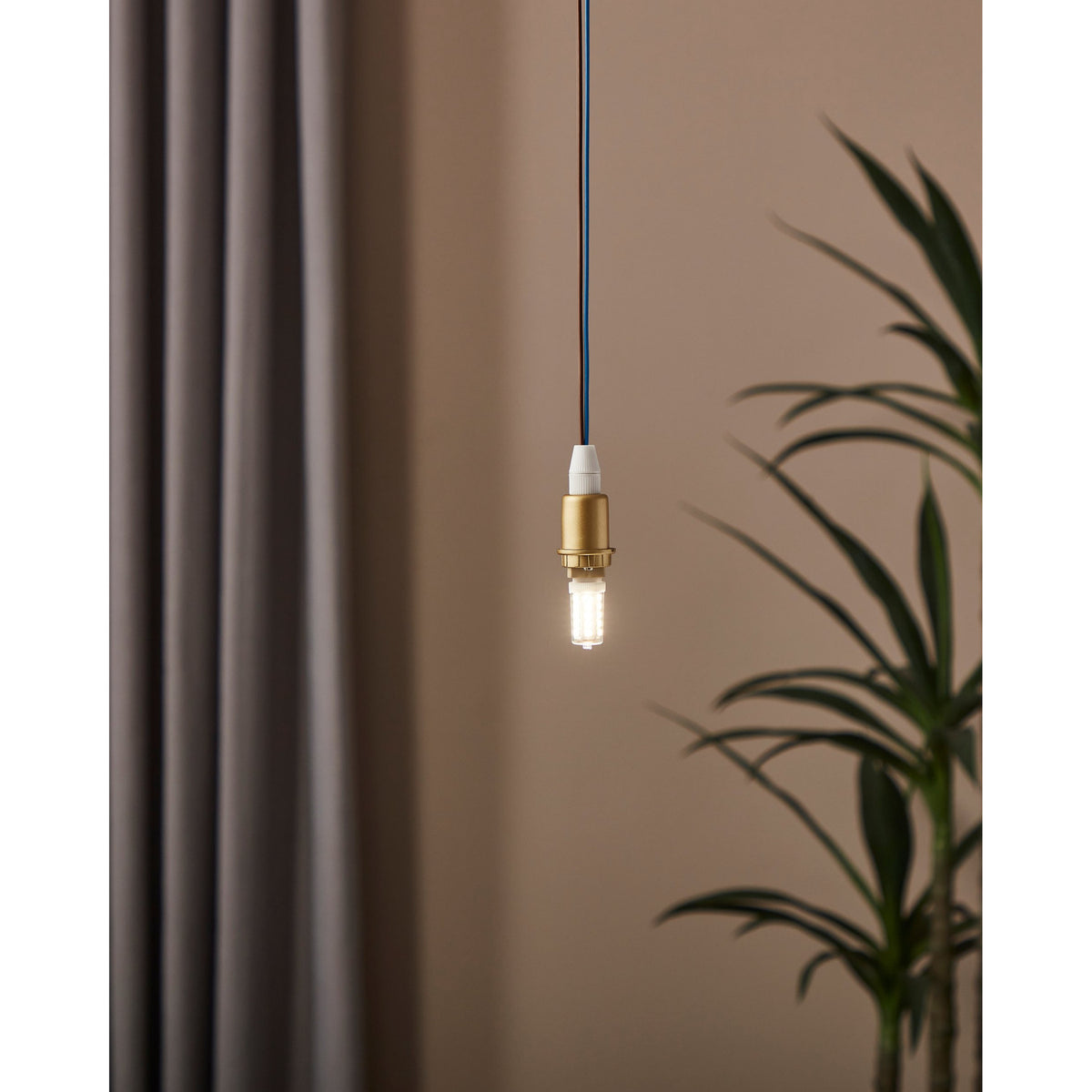 Dimmable LED G9 capsule bulb lit warm white in a brass pendant fitting, shown hanging against a neutral wall beside a curtain and plant.