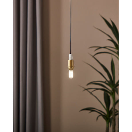 Dimmable LED G9 capsule bulb lit warm white in a brass pendant fitting, shown hanging against a neutral wall beside a curtain and plant.