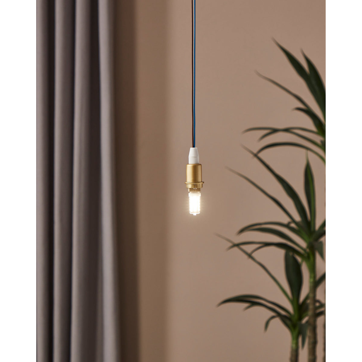 Pendant light fitted with a dimmable G9 LED capsule bulb, brass holder and dark cable, glowing warm white against a neutral wall with curtain and plant nearby.