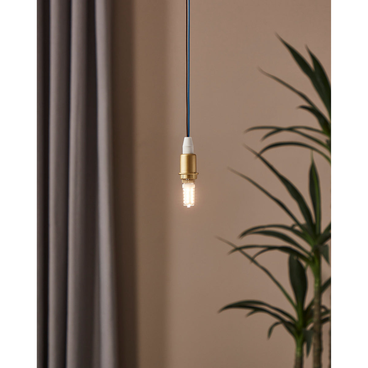 Dimmable LED G9 capsule bulb glowing warm white, hung from a brass-toned pendant fitting against a neutral wall with a curtain and plant nearby.