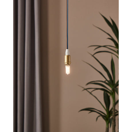 Dimmable LED G9 capsule bulb glowing warm white, hung from a brass-toned pendant fitting against a neutral wall with a curtain and plant nearby.