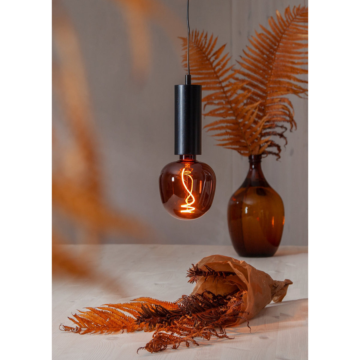 Dimmable LED Globe Bulb E27 G125 in cognac-tinted glass with a spiral filament, shown lit in a black pendant fixture above a table styled with dried ferns and an amber glass vase.