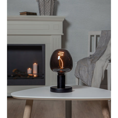 Dimmable LED G125 globe bulb in smoked black glass with a visible spiral filament, fitted to a matte black table lamp base on a coffee table beside a fireplace.
