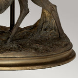 The Amelia-1 Table Lamp with an oval shade showcases a beautifully detailed bronze sculpture of stag's legs and hooves, set on a textured circular base with an exquisite patina, all elegantly contrasted by a neutral gray background.