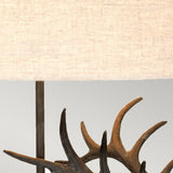 A close-up of the Angus-2 Table Lamp, featuring a beige linen oval shade atop a decorative base shaped like antlers with a bronze patina. These dark brown antlers enhance the rustic and natural aesthetic against a light gray background.