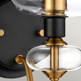 Close-up of the Armand 1 Light Wall Light, showcasing a clear glass orb and a sophisticated aged brass plated and charcoal black paint design. The fixture combines smooth, rounded shapes with charcoal black accents for an elegant appearance.