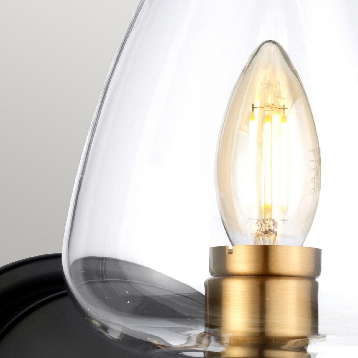 A close-up of the Armand 1 Light Wall Light features a lit, candle-shaped LED bulb encased in a clear glass shade. The bulb's warm glow accentuates its aged brass fitting, beautifully contrasted against a soft gray background.