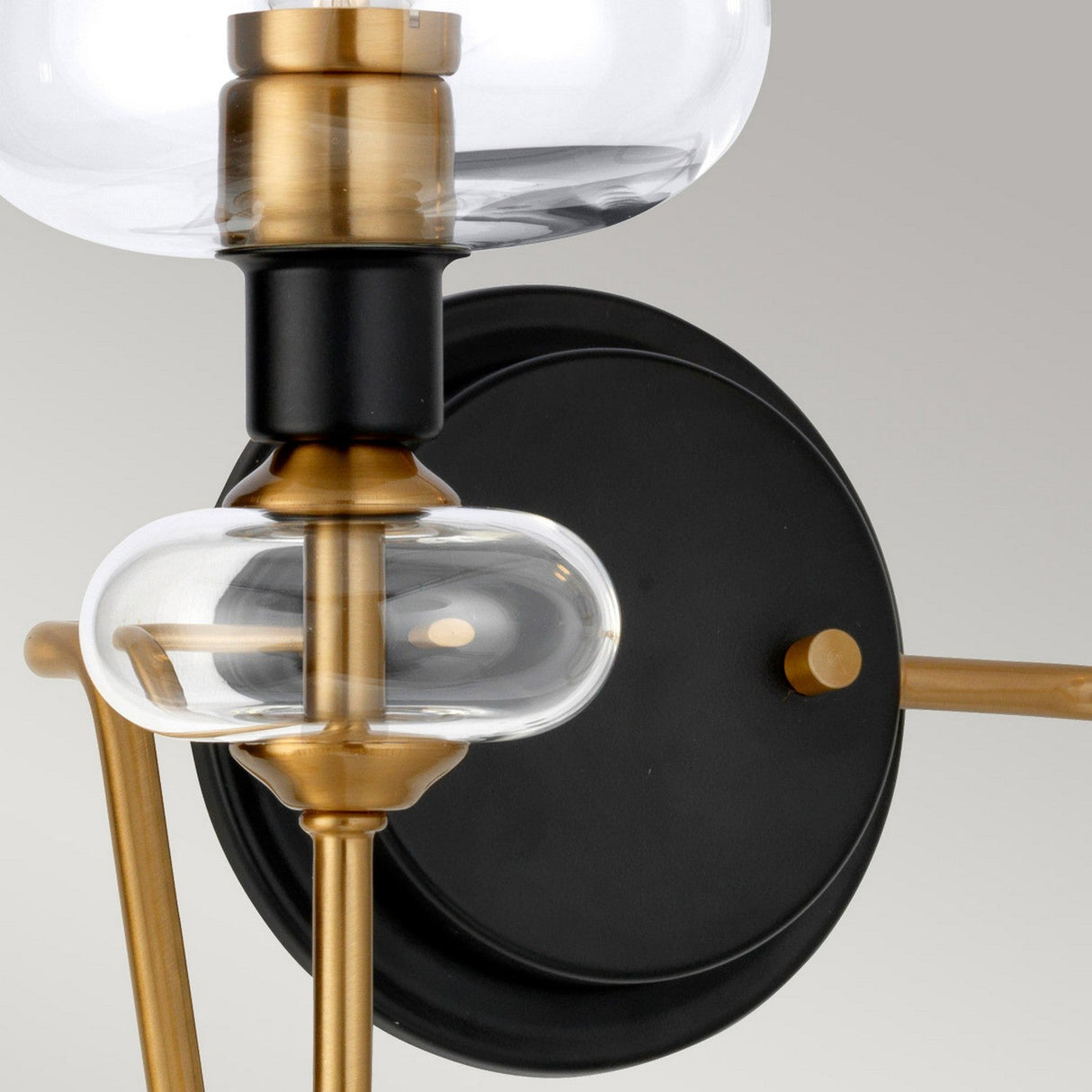 A close-up view of the Armand 2 Light Wall Light, featuring a fashionable charcoal black dual-arm design with an aged brass plated modern circular base. The sophisticated light includes two clear glass elements—a dome-shaped bulb cover and an elongated oval—gracefully attached to radiant gold stems.