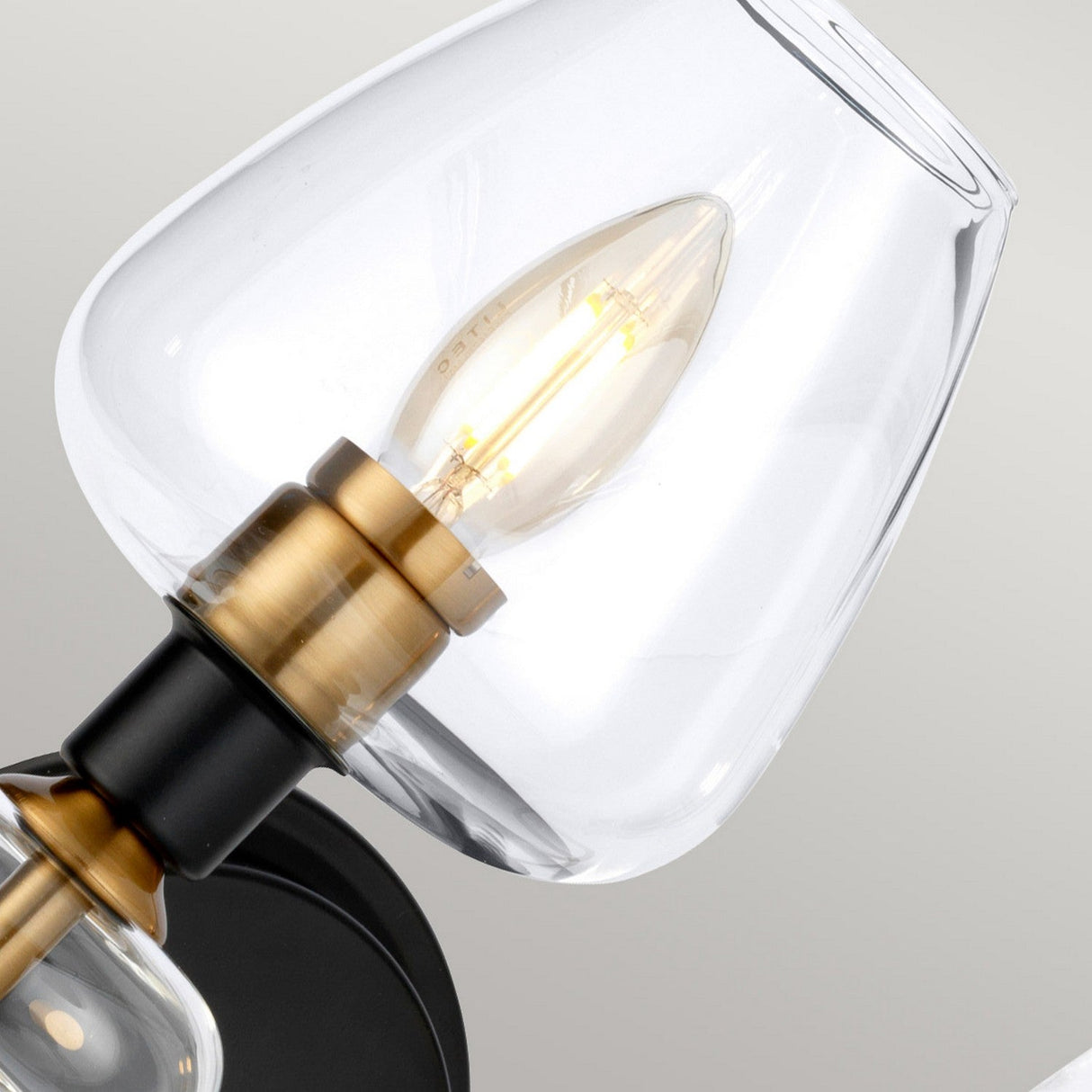 Close-up of the Armand 2 Light Wall Light, showcasing its dual-arm design with a transparent glass shade and an exposed filament bulb. The charcoal black accents seamlessly blend with aged brass-plated details, highlighting its contemporary style against a neutral backdrop.