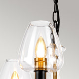 Here's a close-up of the Armand 5 Light Chandelier, designed with modern elegance. It features clear glass shades and emits a warm glow from its bulbs. The fixture is suspended by a charcoal black painted metal chain against a gray backdrop, complemented by aged brass plated accents for an added touch of sophistication.