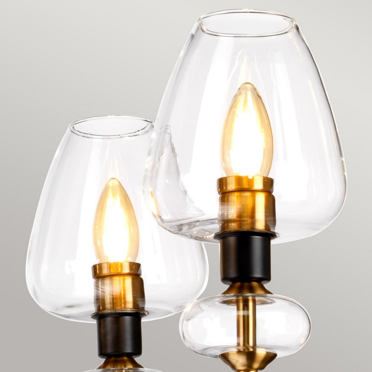 A close-up view showcases two Armand 5 Light Chandeliers, each adorned with clear teardrop-shaped glass covers. The fixtures feature aged brass plated and charcoal black hardware, with visible glowing bulbs that embody a refined modern design set against a neutral background.