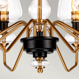 Close-up of the Armand 8 Light Chandelier, showcasing its aged brass plated and charcoal black painted finish. The contemporary design incorporates clear glass elements and multiple symmetrically extending curved arms against a neutral gray background.