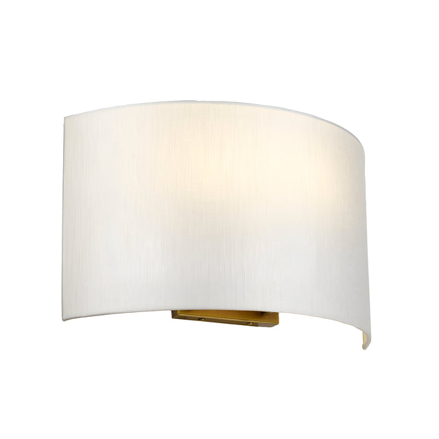 The Cooper Large Curved Wall Light in aged brass, featuring an off-white fabric shade, casts a soft glow against the white background, radiating vintage charm.
