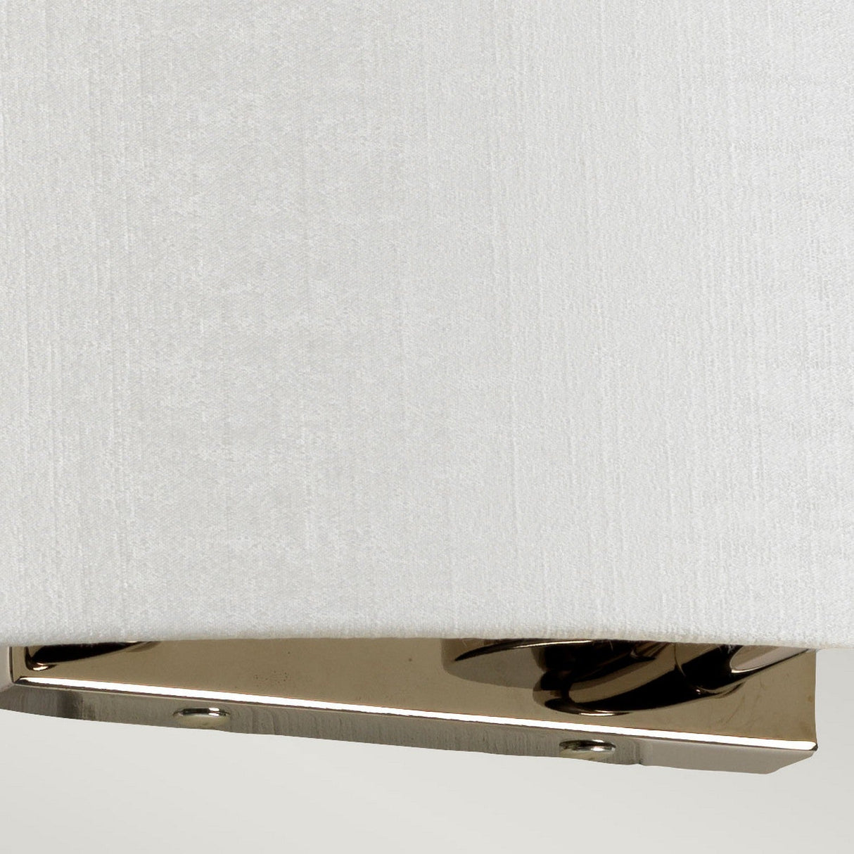 Close-up of the Cooper Large Curved Wall Light featuring a white faux silk lampshade with a polished chrome fixture at the bottom. The shade's fabric has a subtle texture, complementing the shiny, reflective metal surface. A plain, light-colored background enhances its ambient lighting effect.