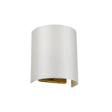 The Cooper Small Curved Wall Light in aged brass features a contemporary design with a curved, cylindrical white fabric shade that provides ideal ambient lighting. This fixture is open at both the top and bottom, with the interior emitting a gentle golden glow.