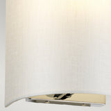 Close-up of the Cooper Small Curved Wall Light, highlighting its compact design with a simple, cylindrical white fabric shade and polished chrome base. This modern wall fixture emits a soft, warm glow that enhances the texture of the fabric.