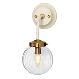 The Cosmos Wall Light - Cream Painted & Aged Brass is a wall-mounted fixture with a cream and brass base. It features a clear ribbed glass globe that houses a visible bulb, blending ambient lighting with retro design. This piece seamlessly combines modern and vintage elements, making it perfect for indoor décor.