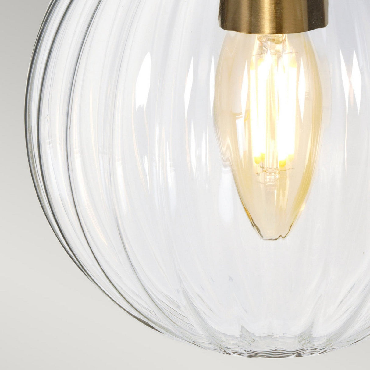 A close-up of the Cosmos Wall Light - Cream Painted & Aged Brass reveals a glowing bulb encased in its clear, ribbed glass fixture, complemented by an aged brass fitting. It emits warm, ambient lighting that fosters a cozy atmosphere against a smooth light gray gradient, exemplifying an exquisite retro design.