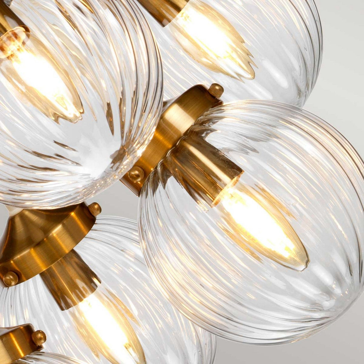 The Cosmos 9 Light Pendant - Cream Painted & Aged Brass features sleek hand-blown glass shades with clear ribbed textures and warm glowing bulbs. Its elegant design includes brass accents and a cream-painted metal structure, making it a sophisticated lighting piece.