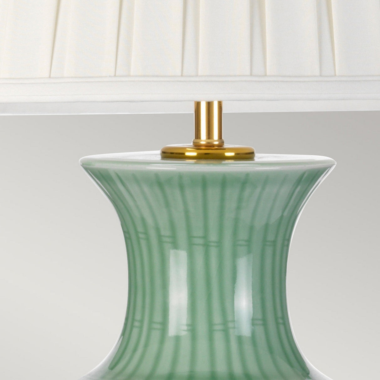 The Dalian Table Lamp with Tall Empire Shade in Celadon features a fluted, green ceramic base that evokes the style of an Oriental Ceramic Vase, adorned with vertical stripes and finished with a gold fitting. It is topped with a pleated white Empire Shade against a plain, neutral background.