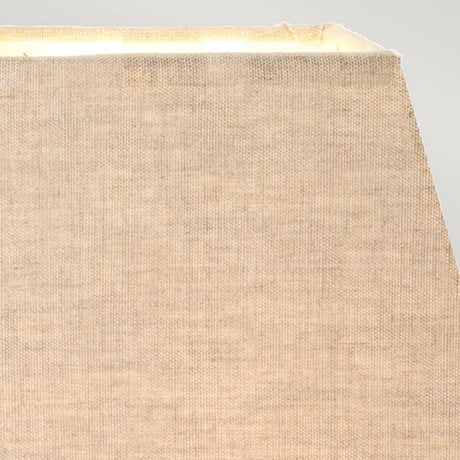 Close-up of a beige, textured fabric lampshade with visible light emanating from the top right, set against a pale gray background. Echoing the Eclipse Table Lamp With Tapered Square Shade - Bronze Patina design, the fabric looks worn, showing some fraying at the edges.